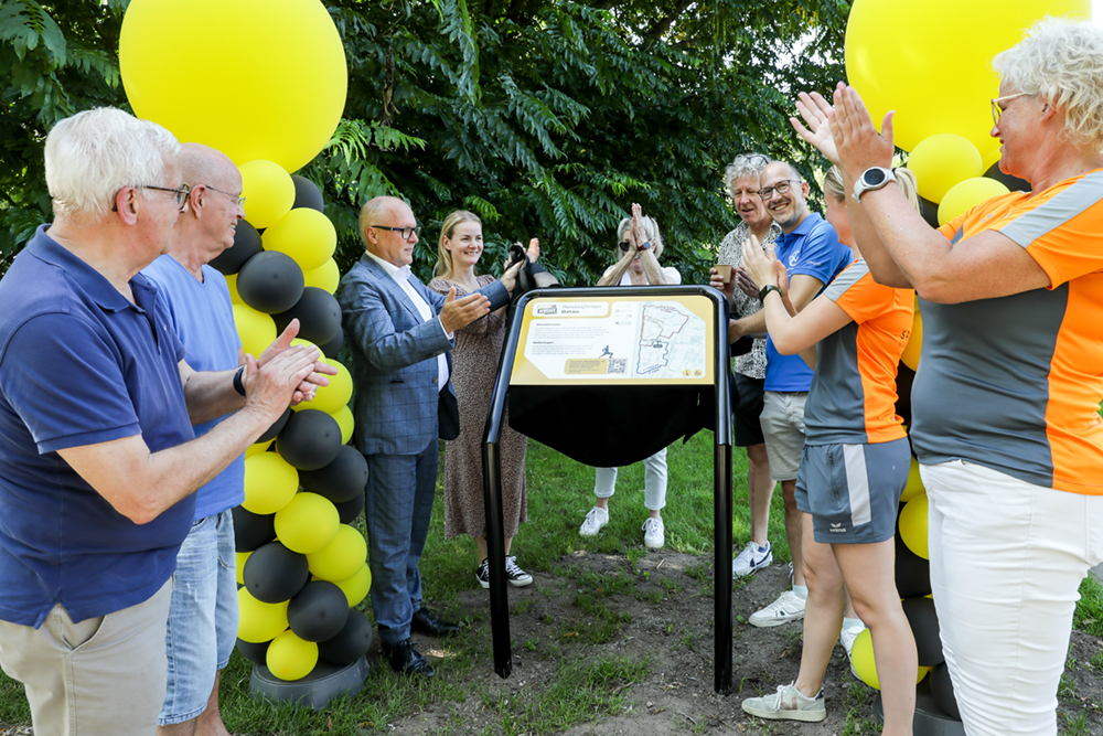 Groepje klappende mensen rondom een informatiebord en gele en zwarte ballonnen